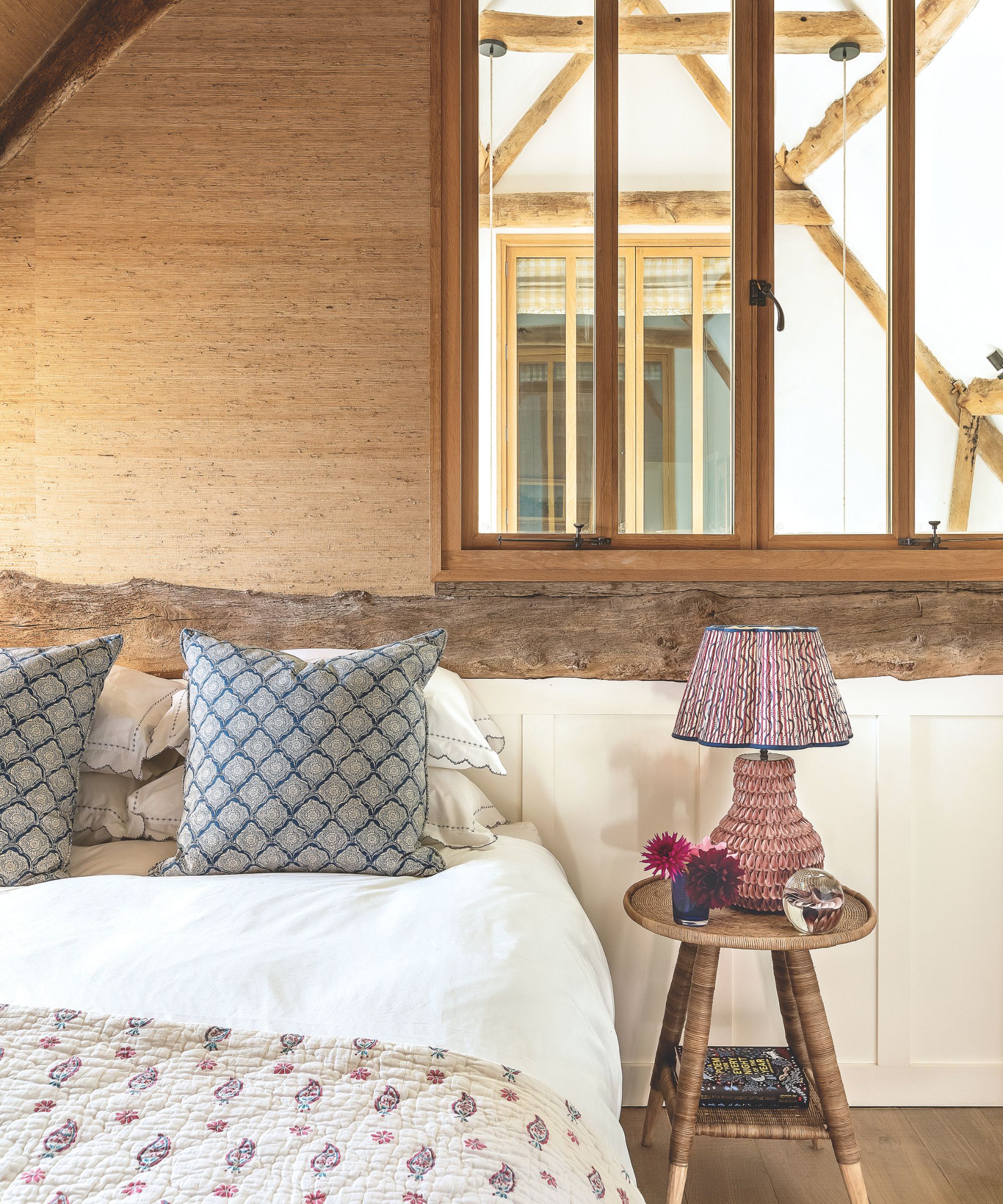 Rustic bedroom with exposed wooden beams. A small wooden bed side table sits next to the bed and the bed in dressed in white sheets with two patterned blue throw pillows and a patterned cream quilt