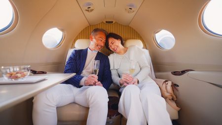 A senior couple is sitting and traveling by a private jet while enjoying champagne and chatting with each other.