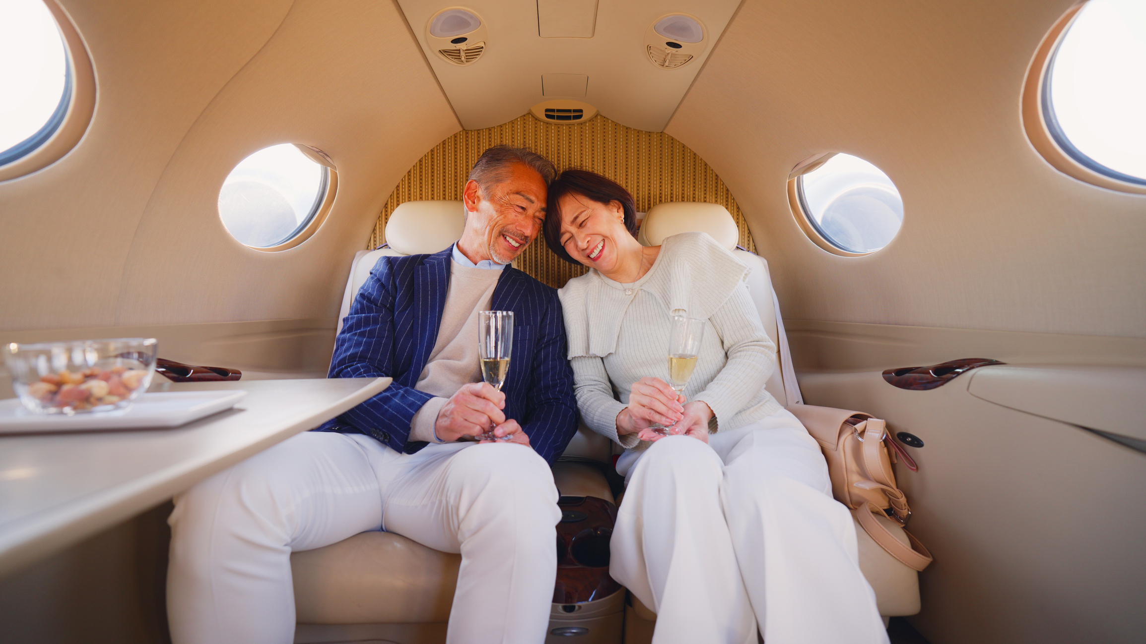 A senior couple is sitting and traveling by a private jet while enjoying champagne and chatting with each other.