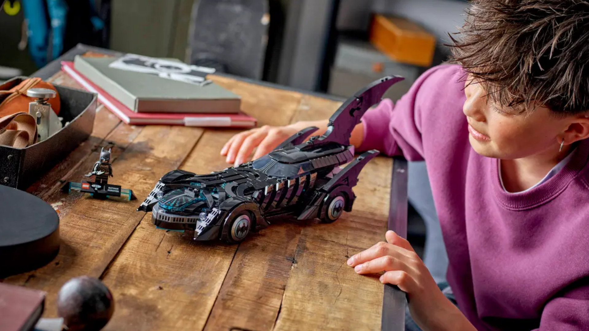 A child smiles at the Lego Batman Forever Batmobile as it sits on a wooden table beside the minifigure, which is on a plaque