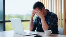 Visibly stressed male office worker with hands pressed against forehead while working on a laptop.