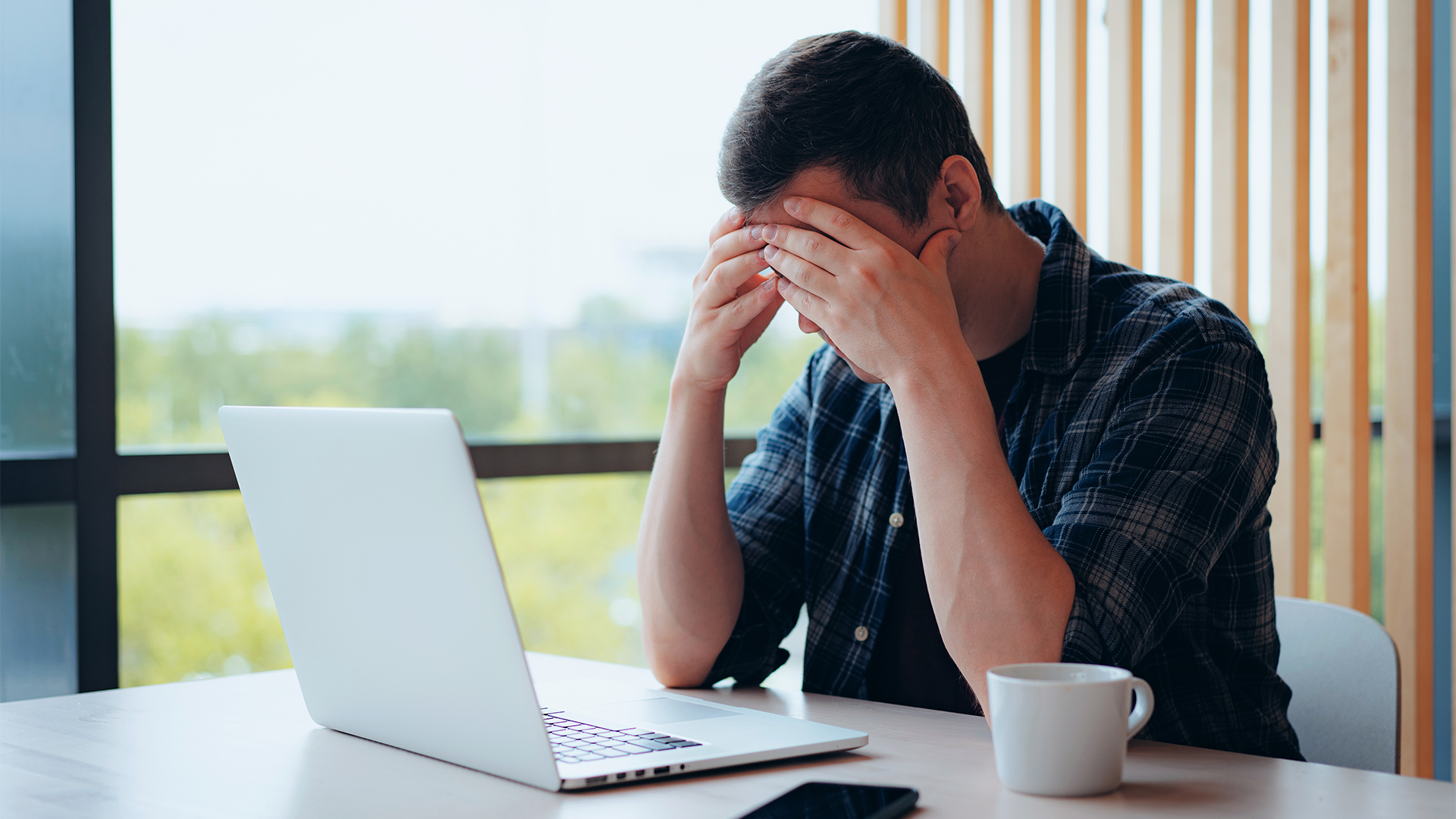 Visibly stressed male office worker with hands pressed against forehead while working on a laptop.