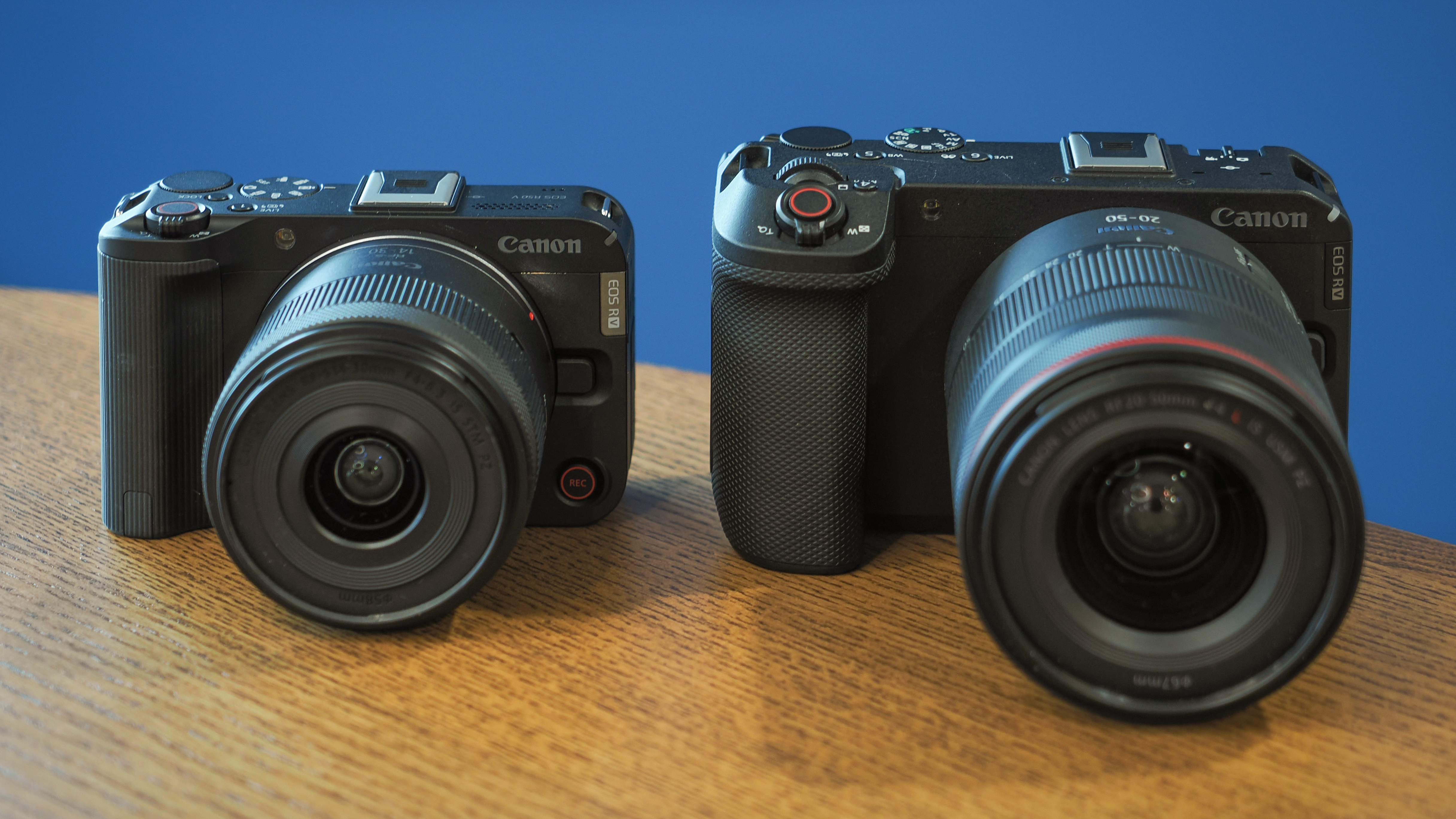 Canon EOS R50 V next to an EOS R6V on a wooden table in front of a blue background