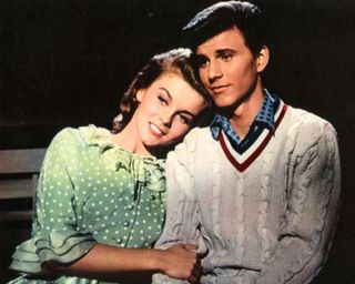 a girl lays her head on a boy's shoulder while sitting on a park bench in Bye bye birdie