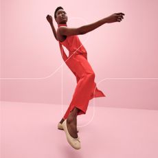 Woman in red jumpsuit posing with beige ballet flats on pink background