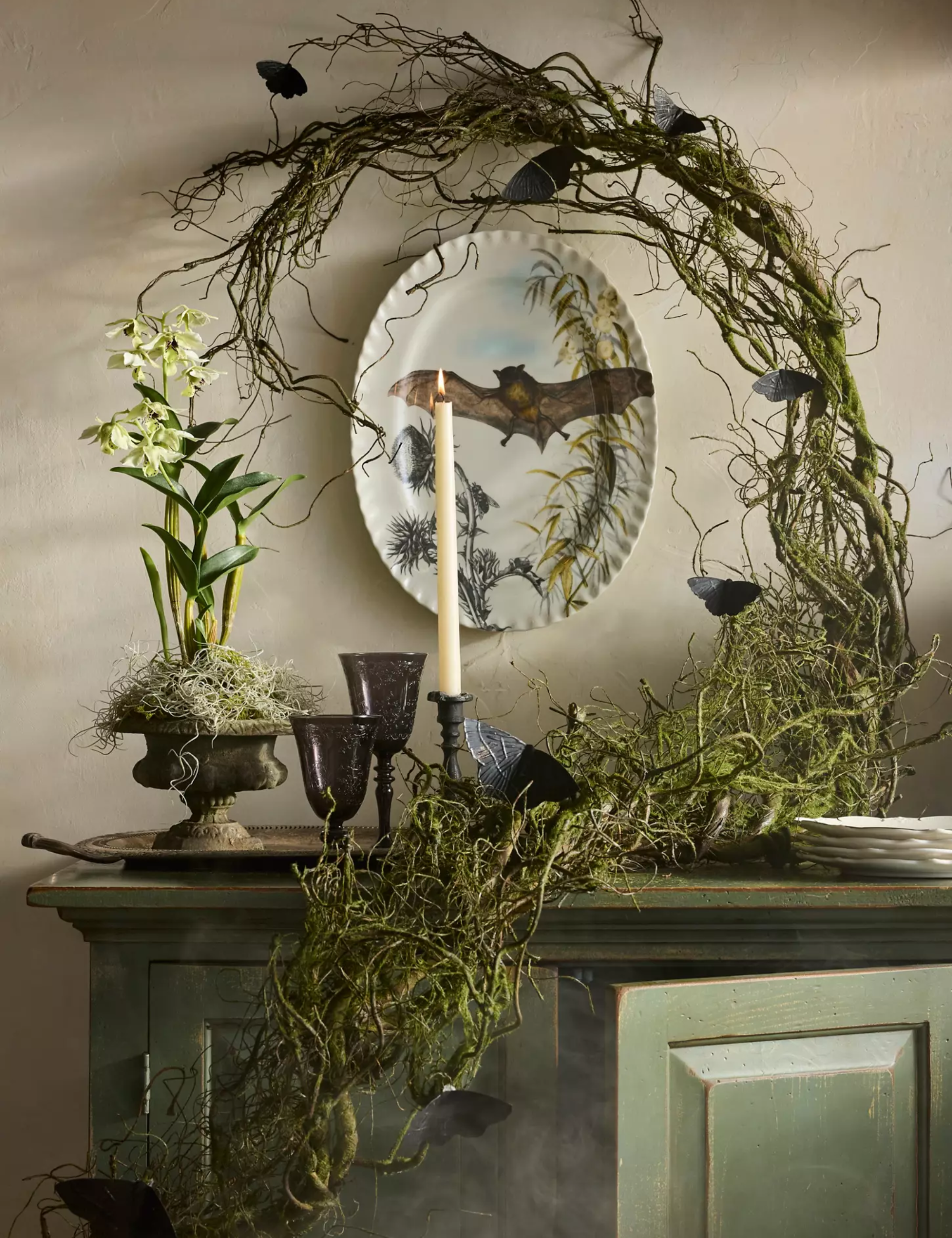 Festive Halloween vignette by Anthropologie featuring faux mossy twig garland, a bat motif porcelain platter, and black couple glasses.