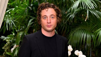 Jeremy Allen White's mirrors are so sleek. Here is the actor wearing a black jumper, standing in front of a dark green leafy background with white flowers
