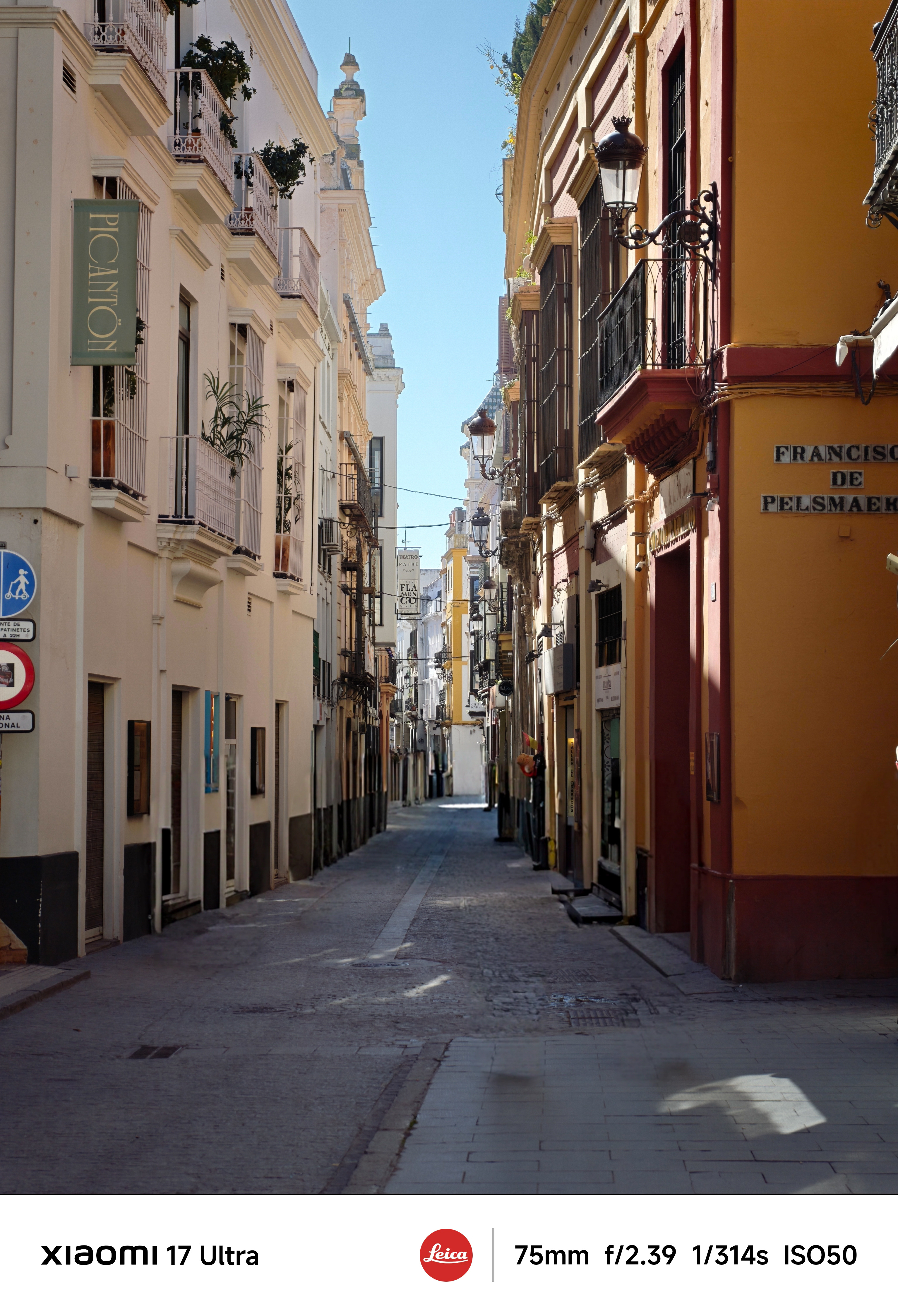 Empty cobbled street framed by cream and ochre buildings with balconies and shop signs under a clear blue sky.