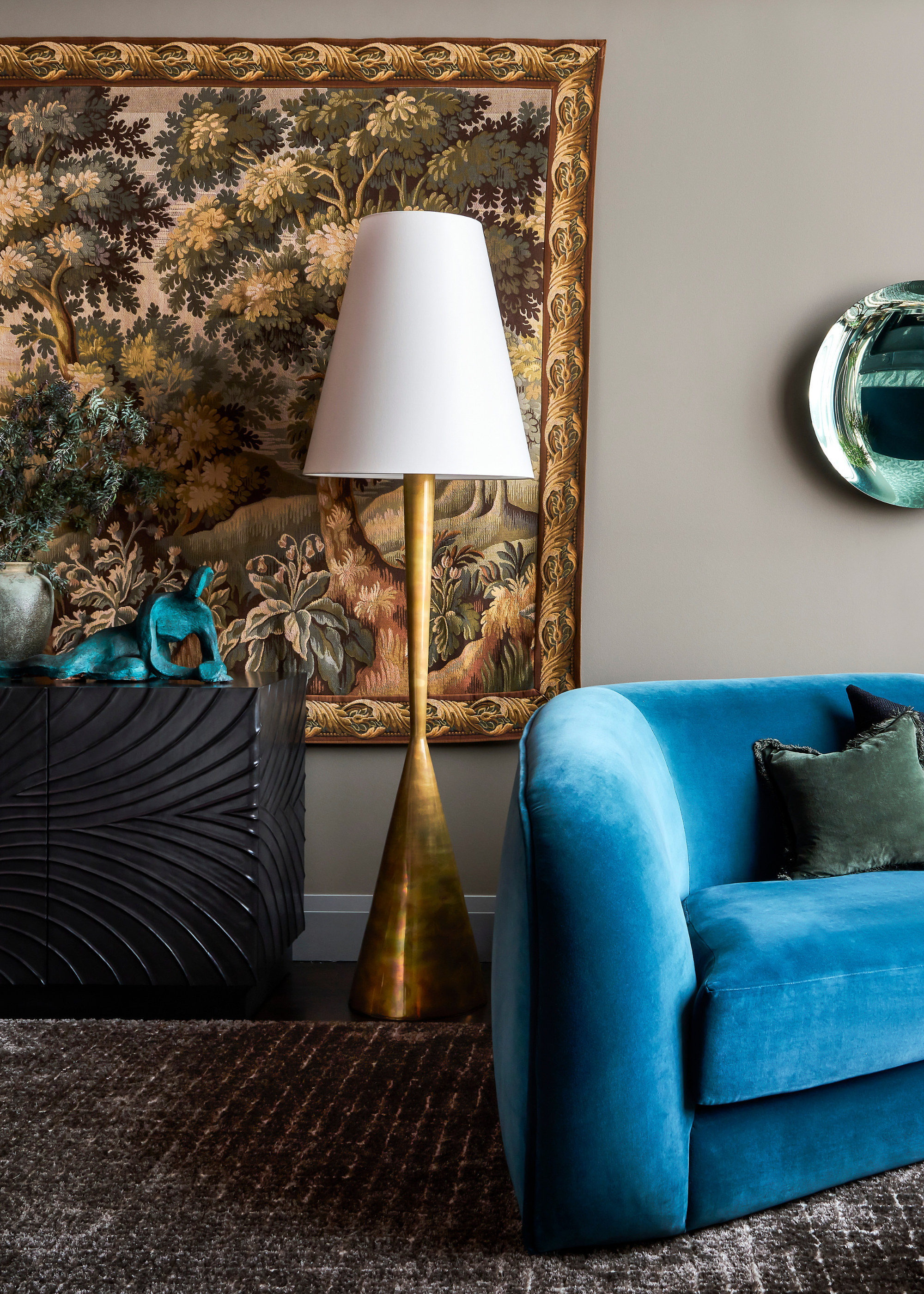 A living room with a black textured sideboard, a large biophilic tapestry with a gold frame on a gray wall, beside a brushed gold standing floor lamp with a white lamp shade, near a rich blue velvet couch and deep green pillows below a reflective turquoise wall accent