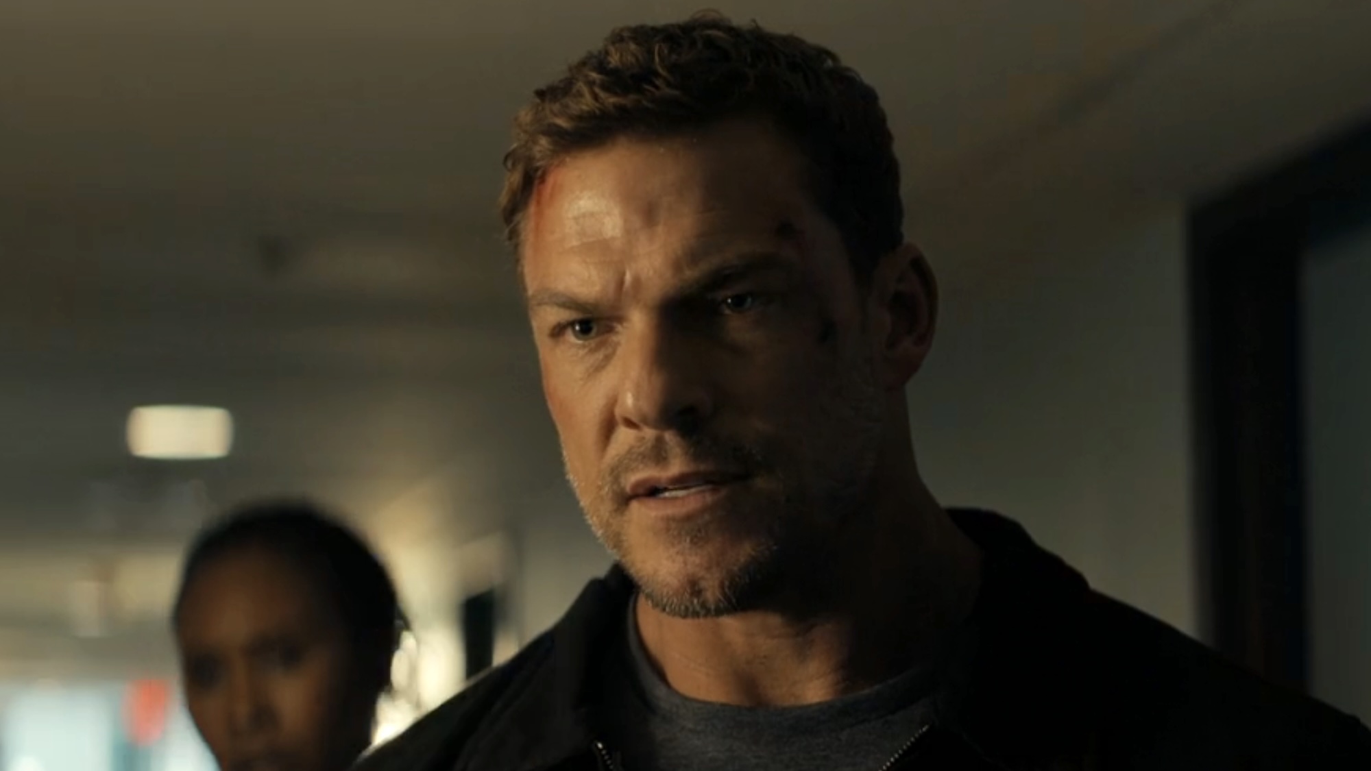 Alan Ritchson's Reacher standing in hotel hallway in a dark maintenance jacket and grey shirt, with scrapes on his forehead in Reacher Season 4