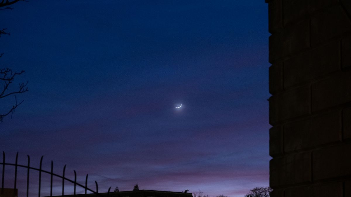 A crescent moon in a purple-and-blue sky, with the silhouette of a fence and a wall on the edges