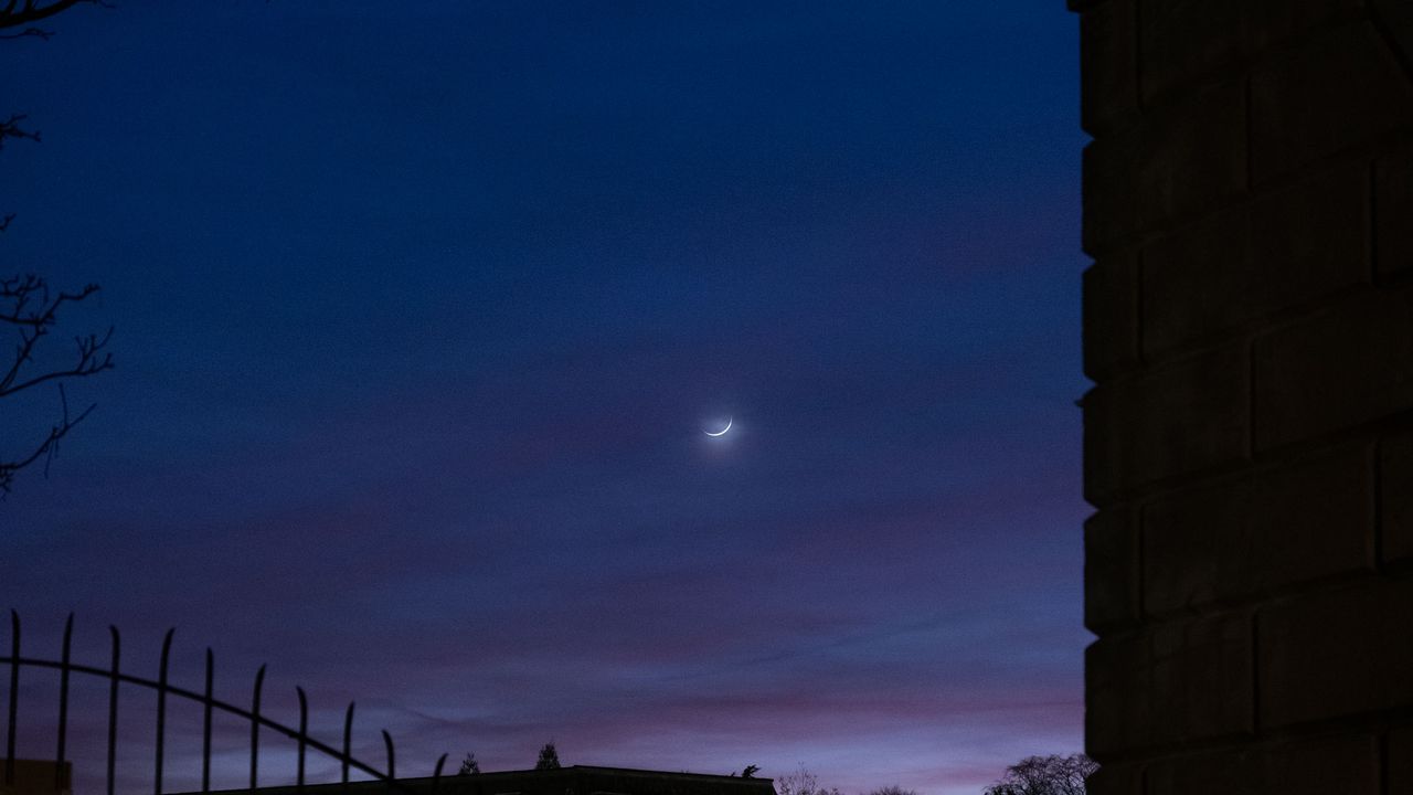 A crescent moon in a purple-and-blue sky, with the silhouette of a fence and a wall on the edges