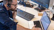 A photo of an IT helpdesk worker speaking to a customer on a headset while sat at their desk and looking at a laptop.