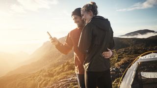 Couple using phone next to tent whilst camping in mountains