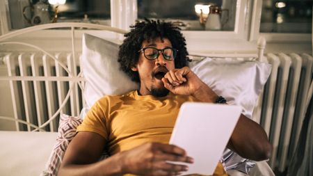 A man lying in bed wearing a yellow t shirt and yawning in the evening as his melatonin levels increase