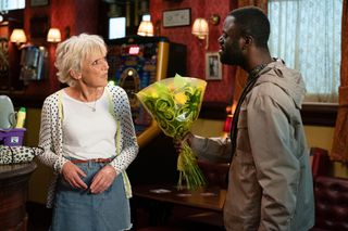 Jean Slater looking at Kojo Asare who is holding a bunch of flowers in the Queen Vic bar