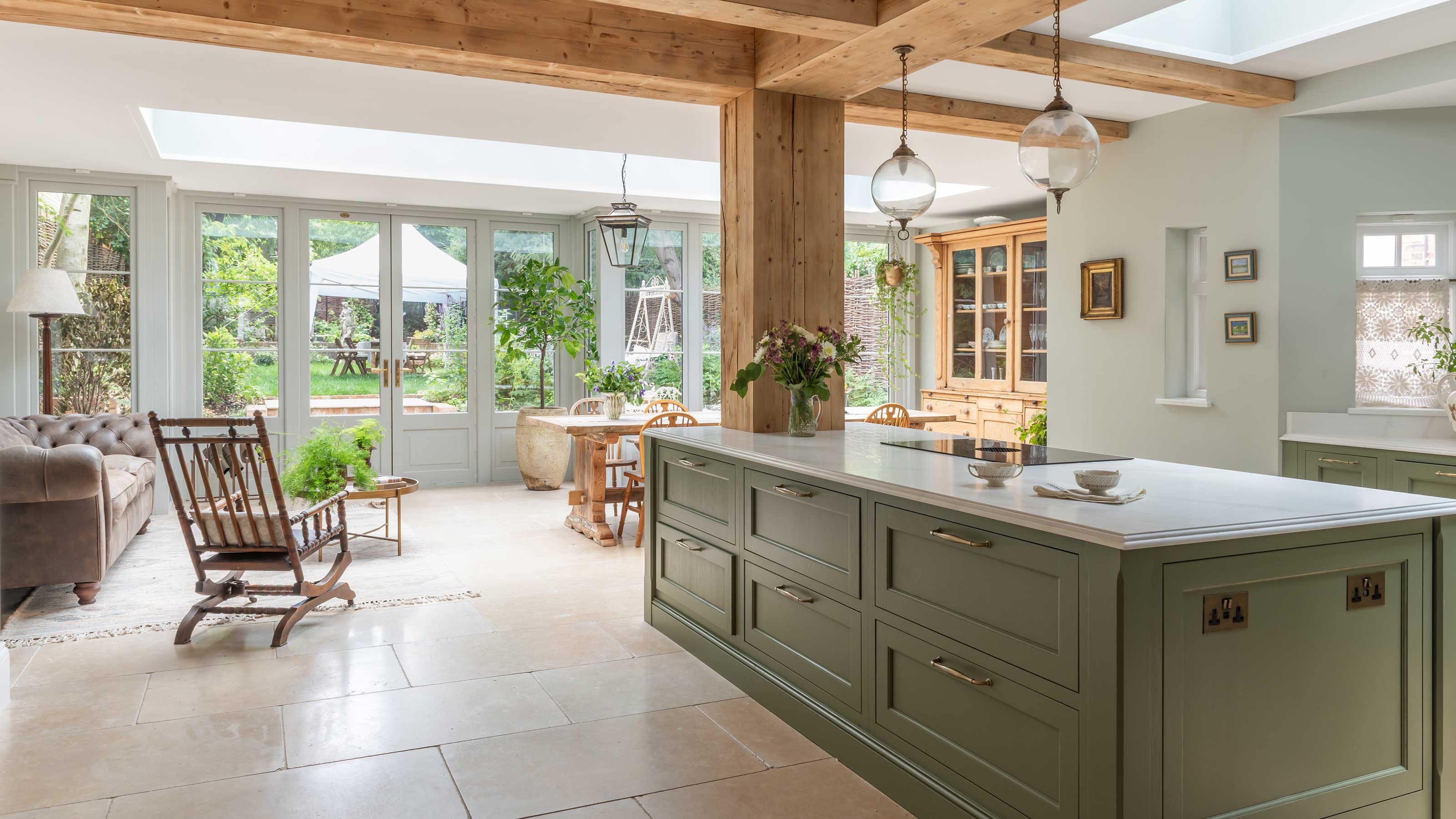 open plan kitchen diner with wooden ceiling and supporting beams, green units, and wall of windows looking out to garden