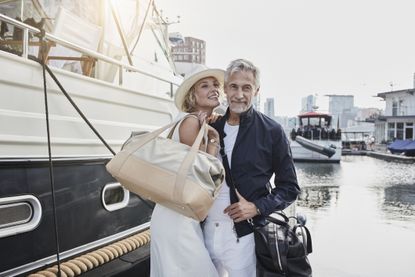 Older man and young woman standing with travelling bags on jetty next to yacht. They look insufferably smug.
