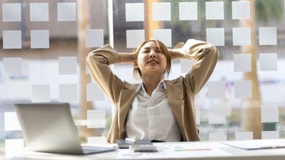 woman wearing a white shirt and beige jacket sitting at a desk with a laptop in front, her hands are behind her head mid-stretch. there's a window behind her and papers on the desk.