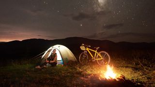 A woman sits by the campfire next to her bike and tent