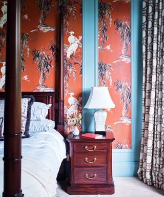 Four poster bed against wallpapered panelled wall wooden bedside table and carpeted floor.