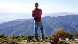 A man wearing jeans stands at the top of a mountain looking at the view with his pack on the ground