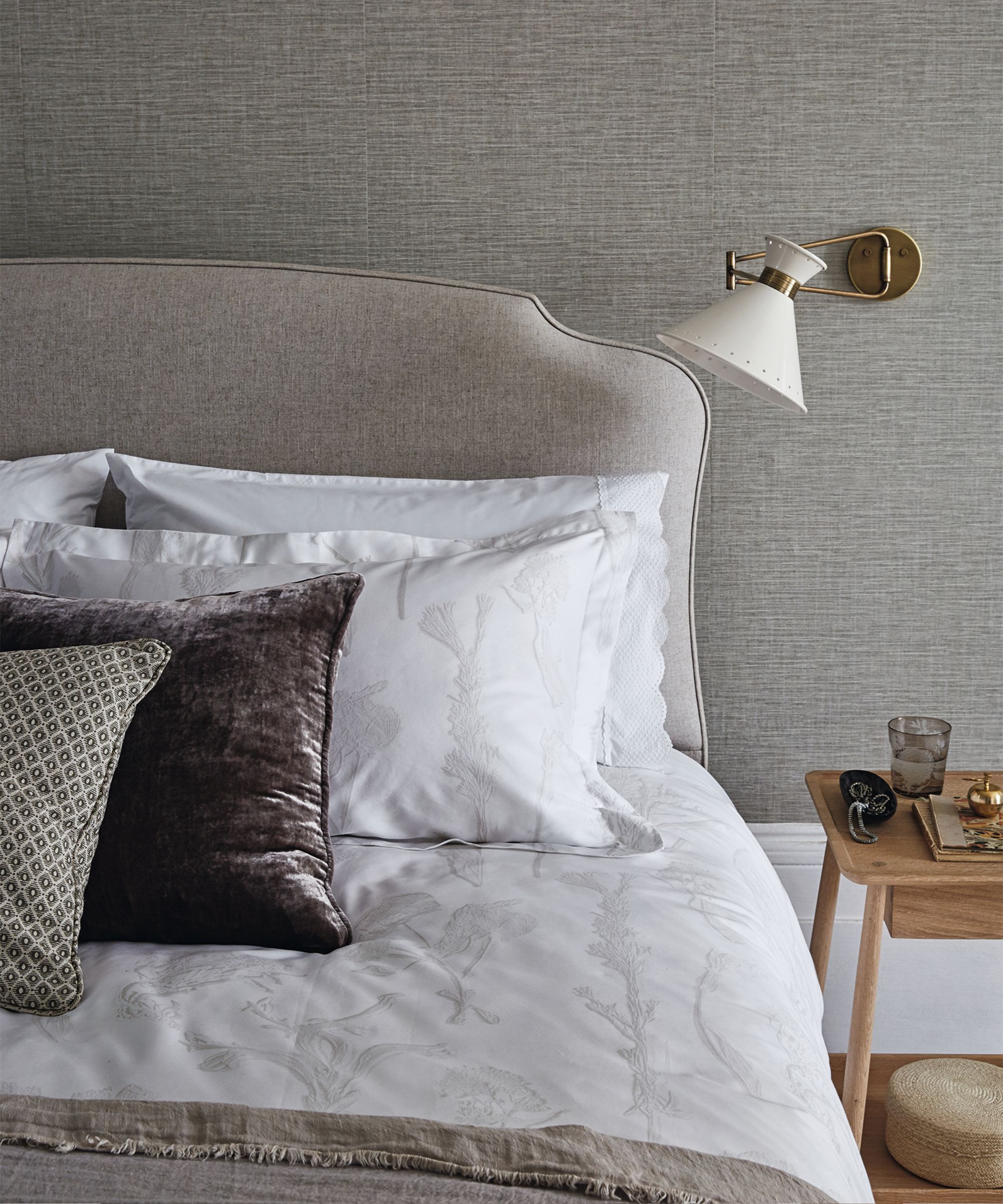 A neutral bedroom space with taupe walls, a grey brown bed with white bedding, assorted cushions, and a wooden bedside table. Behind are white painted baseboards, and the flooring is hardwood.