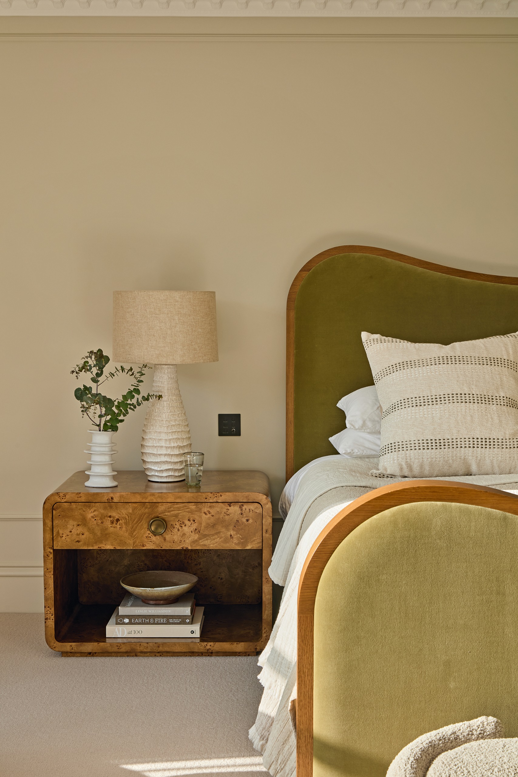Cream bedroom with soft green upholstered bed, burl wood bedside table, cream ceramic bedside lamp and off-white bed linen