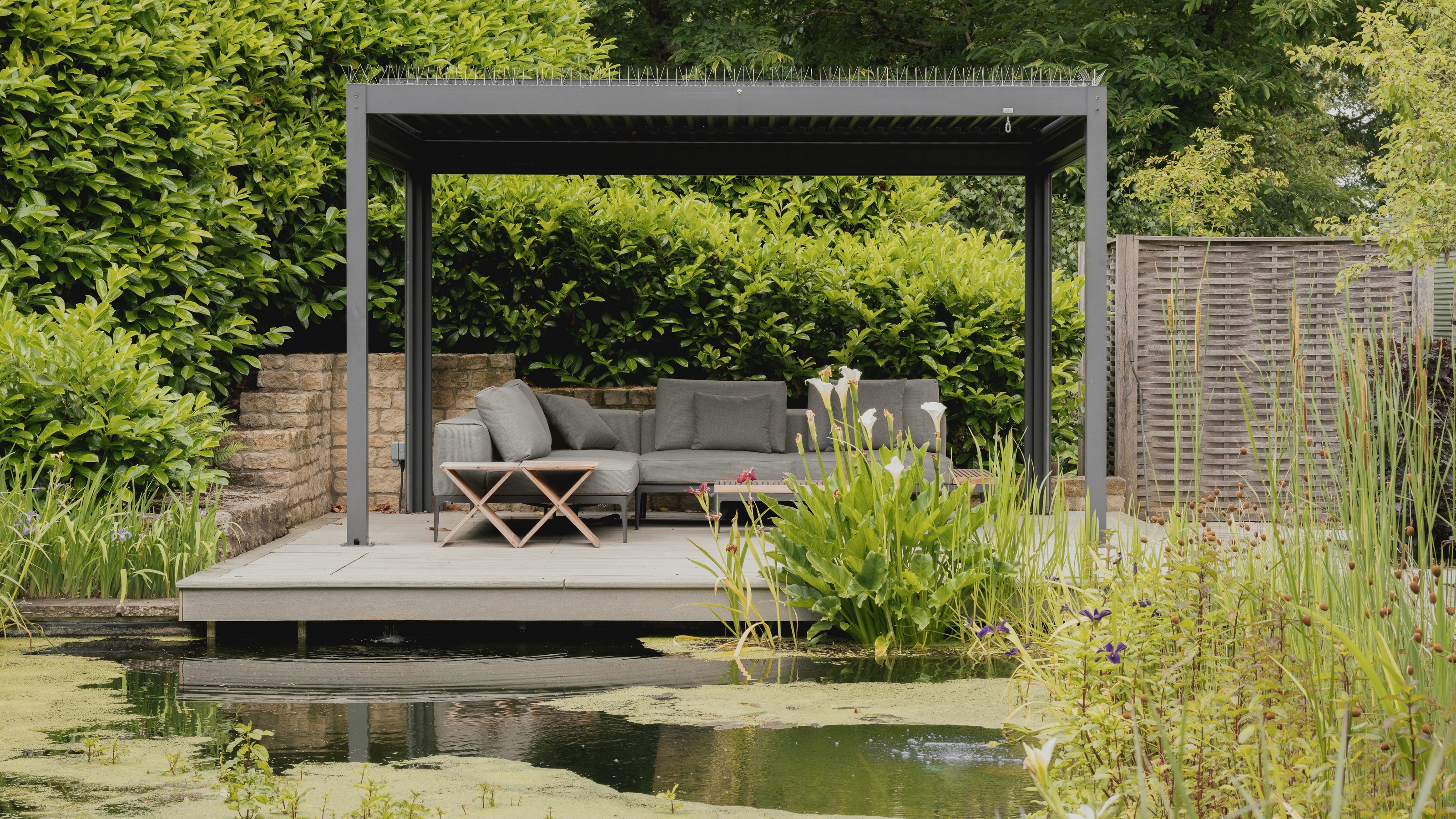 Pergola with retractable roof on a raised platform by a large pond in a garden
