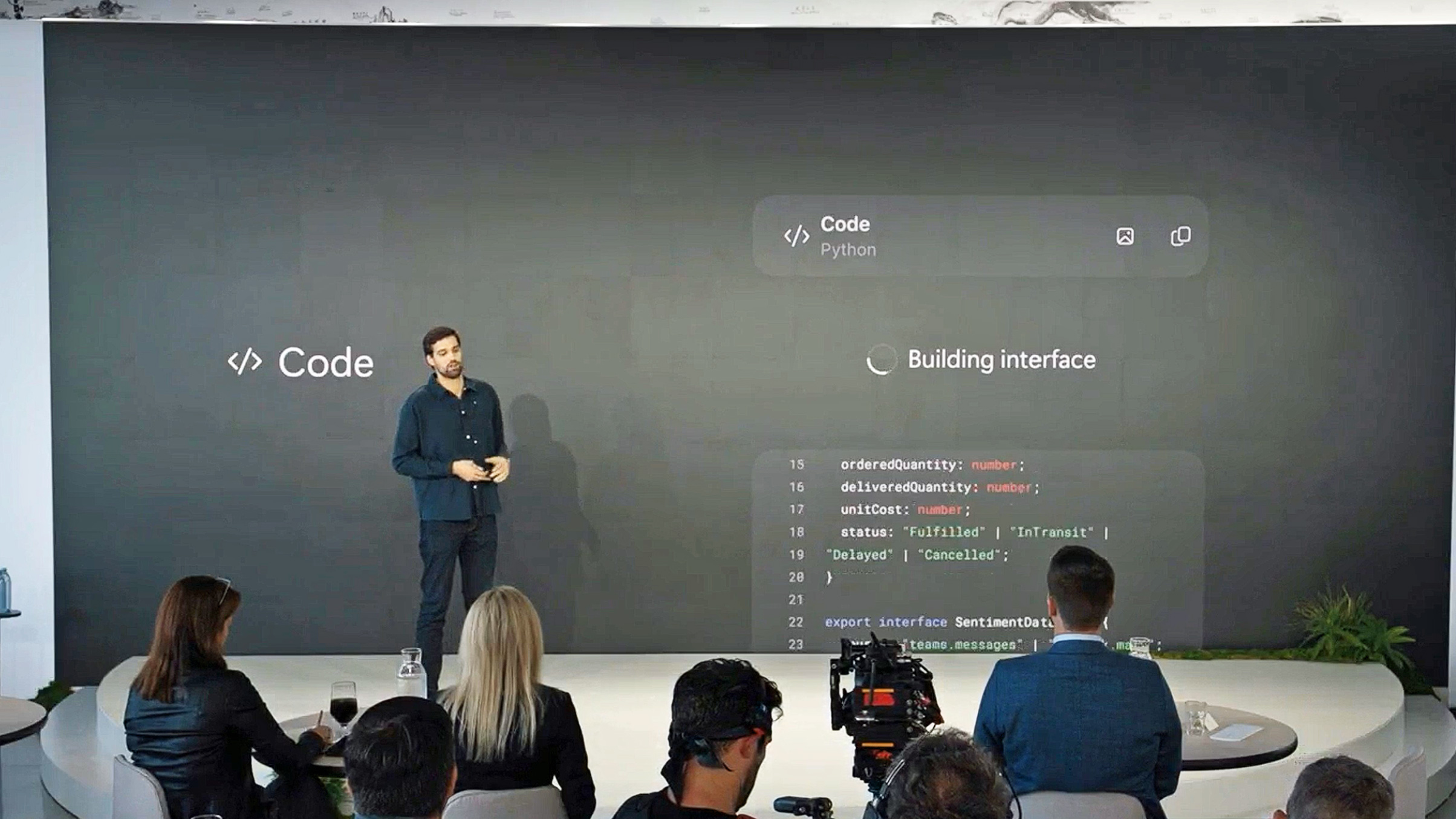 Jacob Andreou stands on stage beside a large screen showing " Code" and live interface code snippets, with an audience and camera equipment in the foreground.