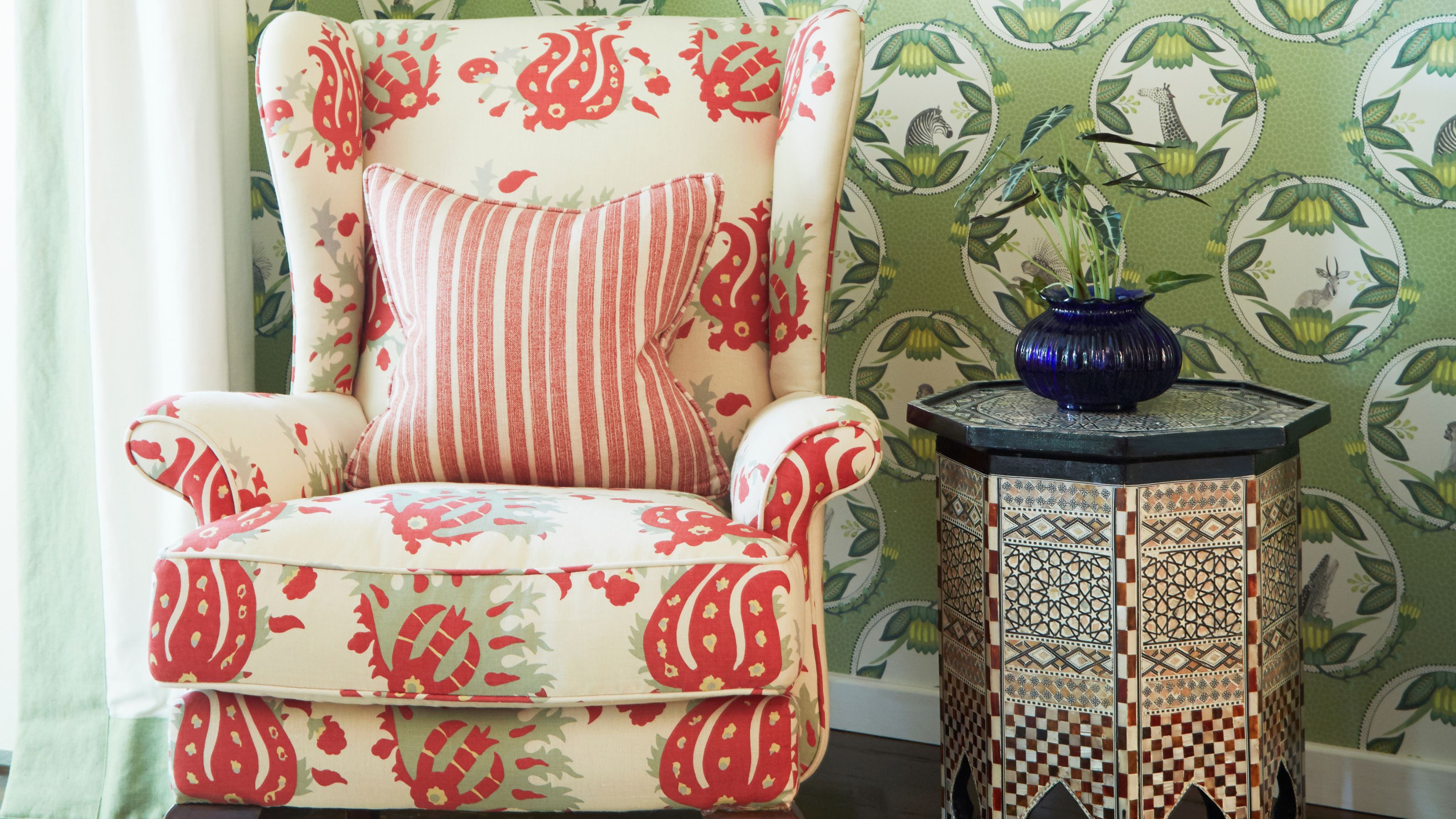 Patterned white and red upholstered chair with mosaic hexagonal side-table with blue glass vase and green plant, and white and green wallpaper behind.