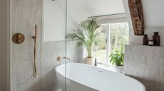 A white tiled bathroom with a white bathtub, gold taps, exposed beams, large window and green leafy houseplants.