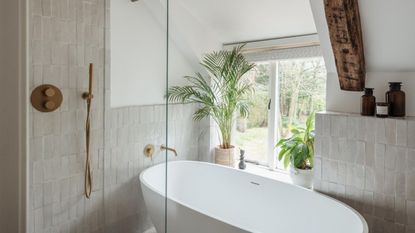 A white tiled bathroom with a white bathtub, gold taps, exposed beams, large window and green leafy houseplants.