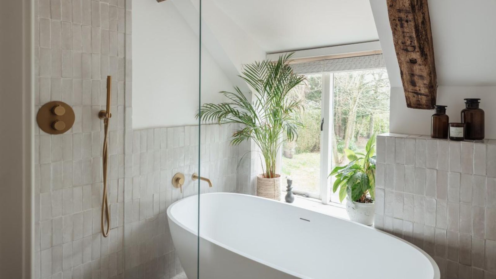 A white tiled bathroom with a white bathtub, gold taps, exposed beams, large window and green leafy houseplants.