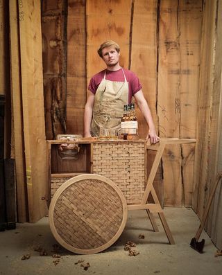 Sebastian Cox standing behind his weaved wood drink trolley. On top of it, there are 3 bottles of The Glenlivet whiskey and two filled glasses.