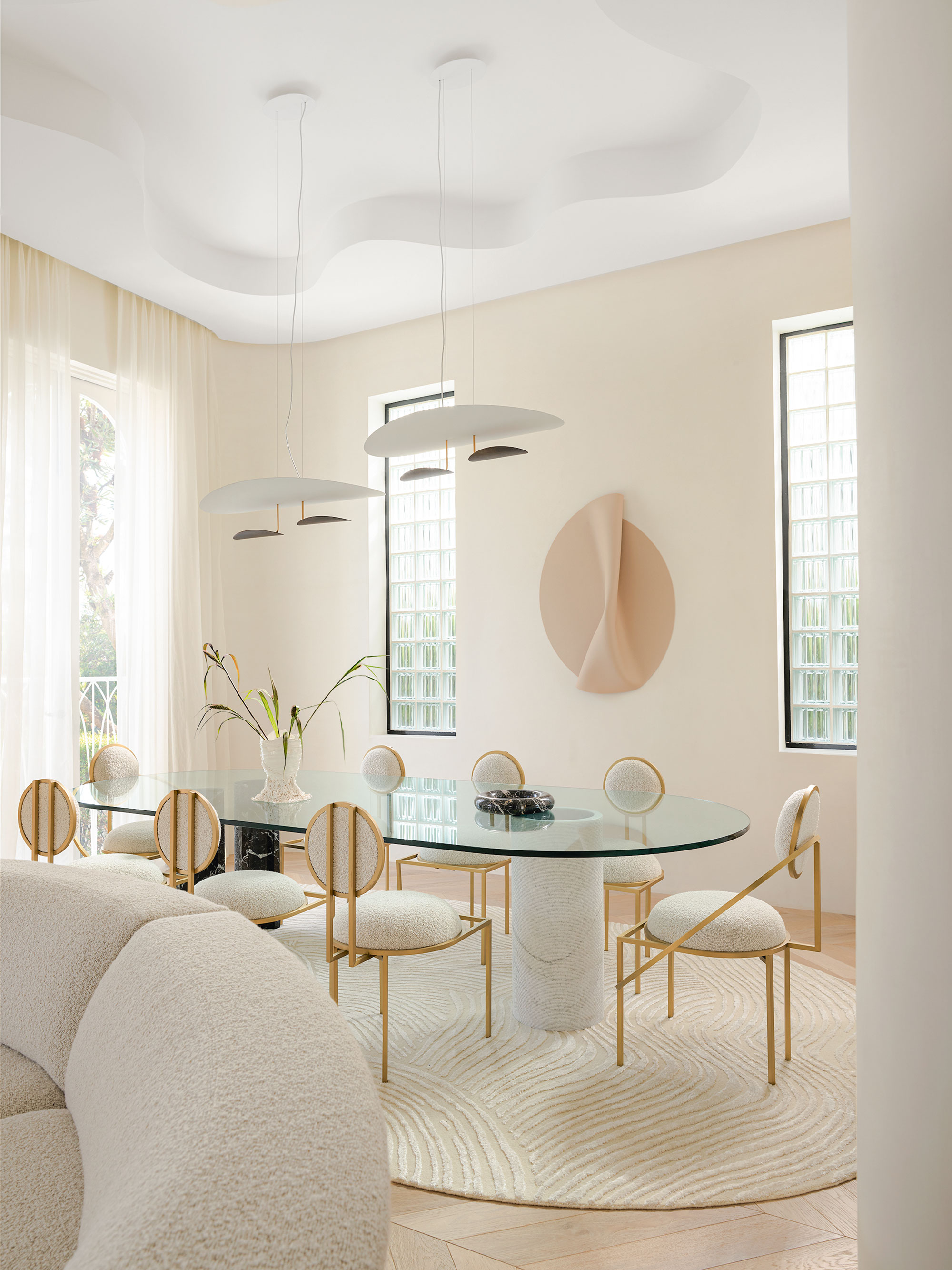 An all cream dining area with two modern pendant lights above the large glass dining table, with all cream fabric and metal dining chairs.