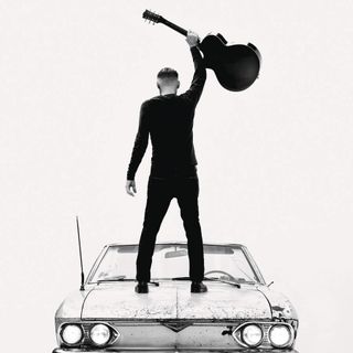 Bryan Adams standing on top of a Chevy Corsair with his back to the camera, holding a guitar aloft.