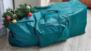 A teal blue duffle-like tree storage bag laying on a wooden floor with a zipped front pocket, for the best Christmas tree storage bags.