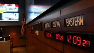 view of digital clocks on brown paneling with a screen far in the background, the screen displays a canadian flag and some blurry images of vessels