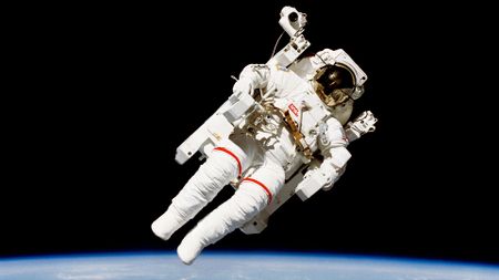 An astronaut wearing a white spacesuit and a jet backpack floats against the blackness of space and the limb of Earth