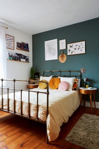 A bedroom with teal feature wall paint decor, yellow and white gingham duvet on bed, framed wall art and floating shelves