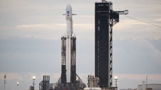a white rocket stands on the launch pad next to its black launch tower, with a gray sky in the background.