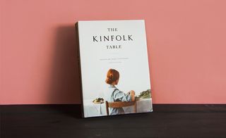 White with black lettering and colour image of a woman sat at a white table with on a wooden chair, fresh vegetables on the top, front cover of the book 'The Kinfolk Table', dark wood surface, orange background