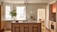 A kitchen with a stone-top island , brass hardware, arched doorways, bright windows, wavy metal stools, and a vase of stems