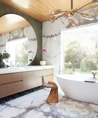 Large bathroom with wooden paneled ceiling, large circular mirror hanging above sink and vanity unit, marble flooring with large white bath, wooden hand seat/decorative object beside bath, geometric glass pendant light above bath
