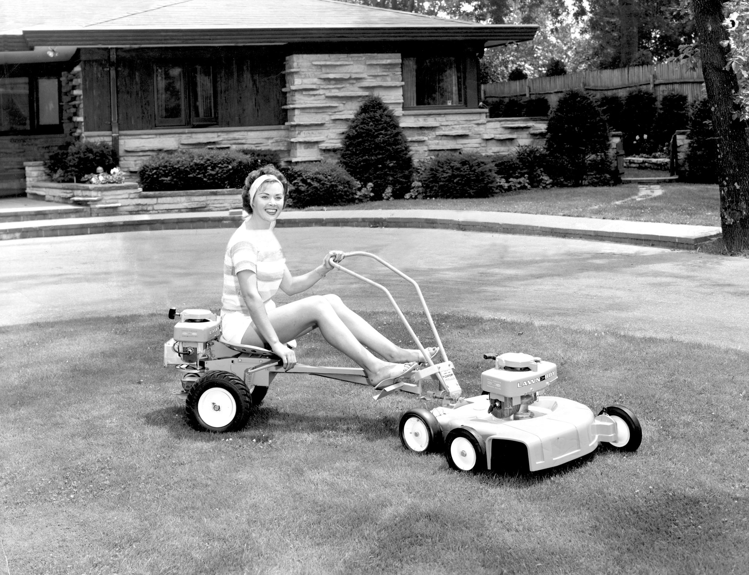 Woman mowing the lawn on a sit down lawnmower. January 1962. 