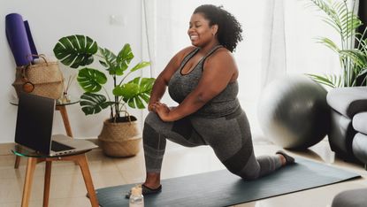 A smiling woman in a sports bra and leggings performs a low lunge in a living room on an exercise mat. Her left knee is on the floor, while her right knee is upright and bent at a 90 degree angle. Both hands rest on her right knee. In front of her is a laptop on a glass table, and in the background we see leafy plants and a yoga mat.