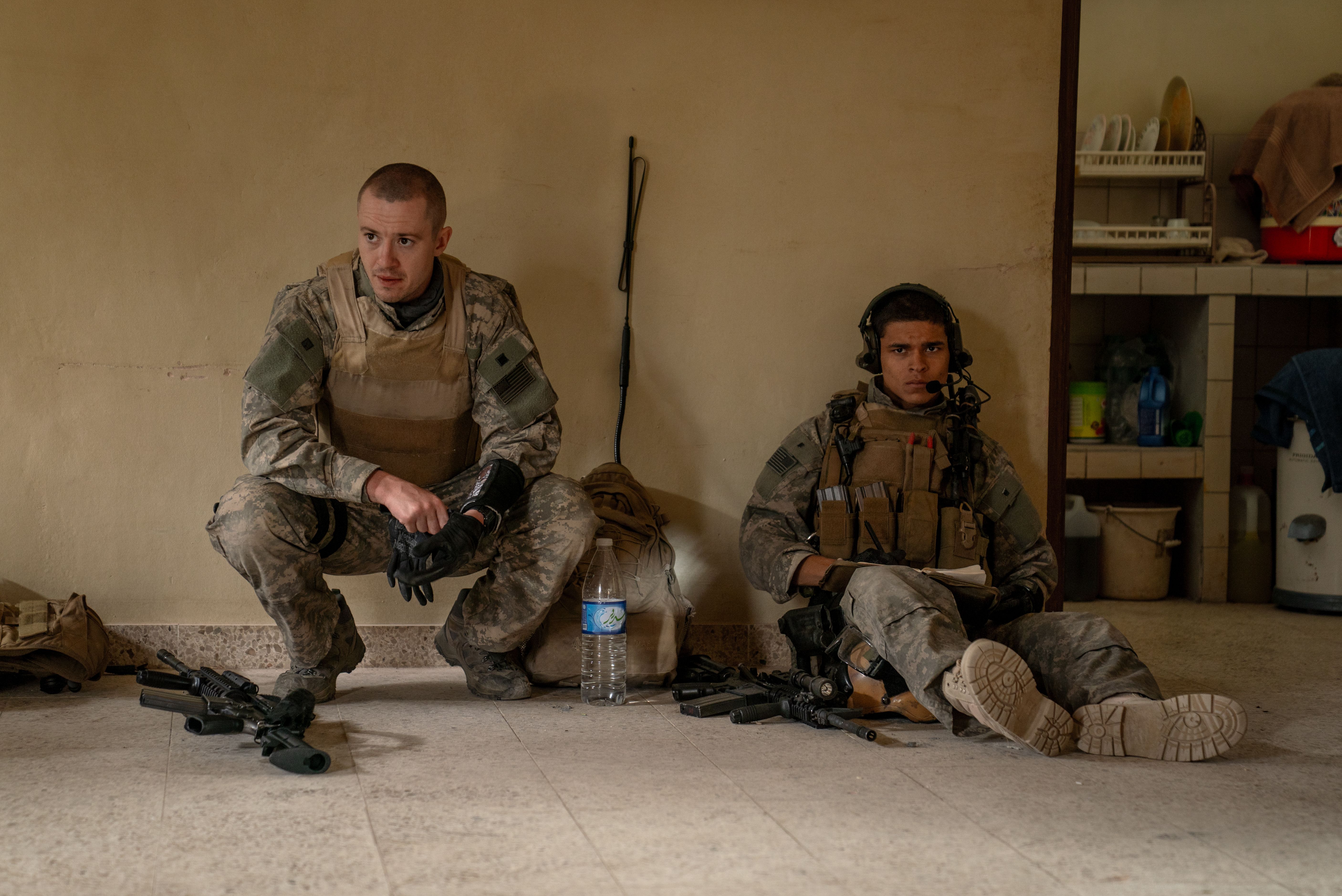 Two soldiers in in full gear in a room with one squatting and one sitting on the floor.