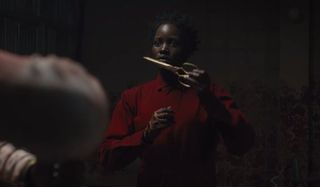 Us Lupita N'yongo holding the scissors in her red jumpsuit, in a darkened room