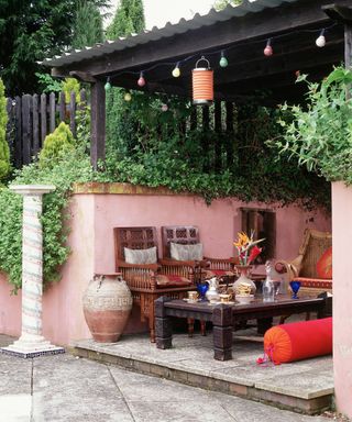 Outdoor patio furniture ideas in Moroccan-inspired woods and red patterned fabrics, in a pink painted alcove with a pergola for shade.